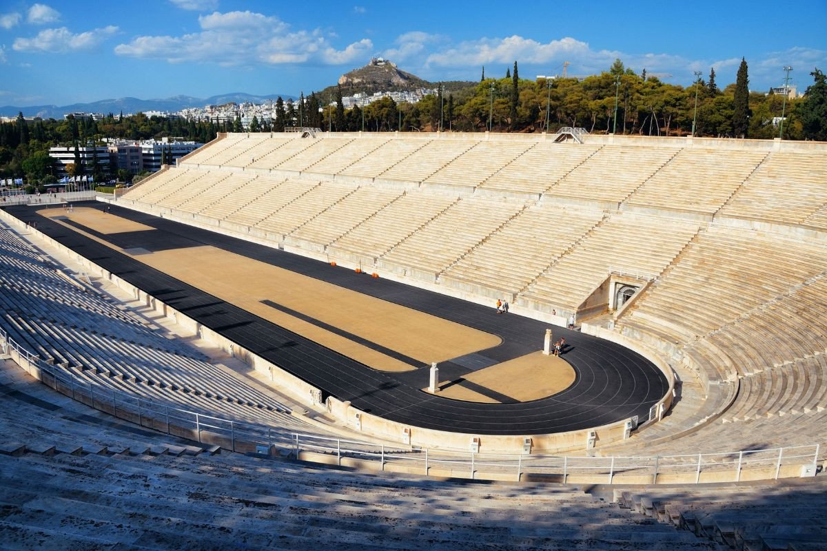 The Panathenaic Stadium in Athens, a large ancient stadium with marble seating, featuring a central running track and surrounded by a backdrop of trees and hills.